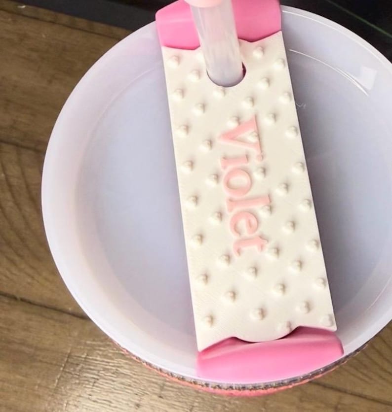 K&ouml;nnte beinhalten: Eine wei&szlig;e und rosa Badebombenform mit dem Wort "Violet" in Rosa. Die Form hat eine strukturierte Oberfl&auml;che mit kleinen, erhabenen Punkten. Die Form ist teilweise in einer wei&szlig;en Sch&uuml;ssel versenkt.