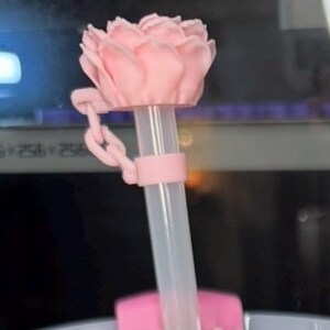 K&ouml;nnte beinhalten: Ein rosa, bl&uuml;tenf&ouml;rmiger Flaschenverschluss mit einem durchsichtigen Strohhalm. Der Verschluss hat ein Kettendetail und einen Ring um den Strohhalm. Die Blume ist hellrosa.