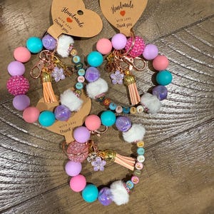 May include: Handmade beaded keychains in pastel colors. Each keychain features a mix of pink, purple, teal, and white beads, along with a tassel, flower charm, and a heart-shaped tag that says "Handmade with love, Thank you."