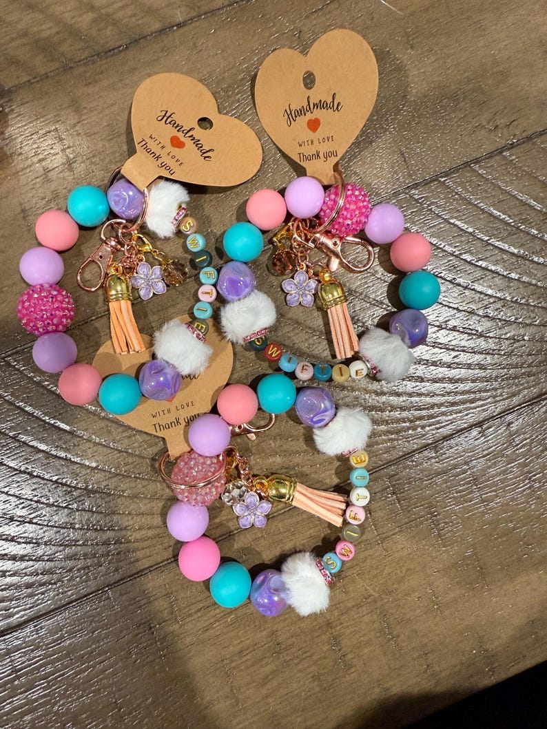 May include: Handmade beaded keychains in pastel colors. Each keychain features a mix of pink, purple, teal, and white beads, along with a tassel, flower charm, and a heart-shaped tag that says "Handmade with love, Thank you."