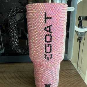 Puede incluir: Vaso rosa cubierto de pedrer&iacute;a iridiscente. La palabra "GOAT" est&aacute; escrita con pedrer&iacute;a negra en el lateral. El asa es negra y est&aacute; cubierta de pedrer&iacute;a. Una cabra de pedrer&iacute;a negra en la parte inferior.
