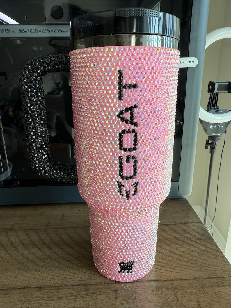 Puede incluir: Vaso rosa cubierto de pedrer&iacute;a iridiscente. La palabra "GOAT" est&aacute; escrita con pedrer&iacute;a negra en el lateral. El asa es negra y est&aacute; cubierta de pedrer&iacute;a. Una cabra de pedrer&iacute;a negra en la parte inferior.