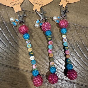 May include: Three handmade keychains with the word "SWIFTIE" spelled out in colorful beads. Each keychain features a pink, sparkly ball, blue and green beads, and a silver clasp. The keychains have heart-shaped tags that read "Handmade with love, Thank you."