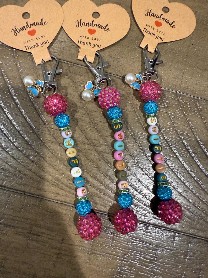 May include: Three handmade keychains with the word "SWIFTIE" spelled out in colorful beads. Each keychain features a pink, sparkly ball, blue and green beads, and a silver clasp. The keychains have heart-shaped tags that read "Handmade with love, Thank you."