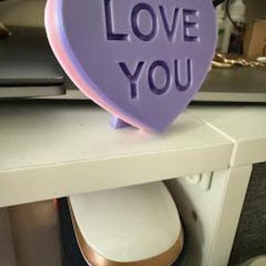 May include: A stacked heart-shaped decoration in shades of purple and pink, with the words "LOVE YOU" carved into the front. The decoration is standing on a white shelf, with a computer mouse visible below.