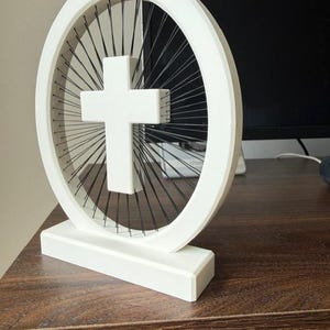 May include: A white, three-dimensional cross sculpture. The cross is centered within a white circular frame, with black strings radiating from the center to the frame. The sculpture sits on a white rectangular base. The background includes a dark monitor and a wooden surface.