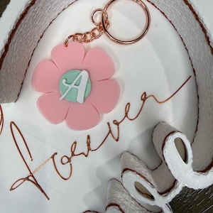 May include: A pink flower-shaped keychain with a rose gold keyring and chain. The flower has a mint green center with the letter "A". The word "forever" is written in rose gold wire on a white background.