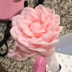 K&ouml;nnte beinhalten: Ein hellrosa, bl&uuml;tenf&ouml;rmiges Objekt mit geschichteten Bl&uuml;tenbl&auml;ttern. Das Objekt ist an einer wei&szlig;-rosa Basis befestigt. Der Hintergrund ist eine gesprenkelte graue Oberfl&auml;che und ein rosa Objekt.