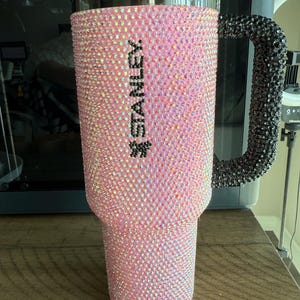 Puede incluir: Un vaso Stanley rosa cubierto de pedrer&iacute;a iridiscente. El asa es negra y tambi&eacute;n est&aacute; cubierta de pedrer&iacute;a. La palabra "STANLEY" est&aacute; impresa verticalmente en negro en el lateral del vaso.