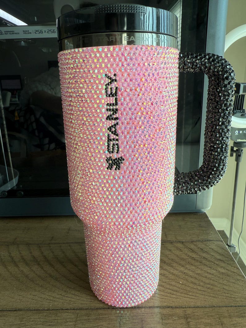 Puede incluir: Un vaso Stanley rosa cubierto de pedrer&iacute;a iridiscente. El asa es negra y tambi&eacute;n est&aacute; cubierta de pedrer&iacute;a. La palabra "STANLEY" est&aacute; impresa verticalmente en negro en el lateral del vaso.