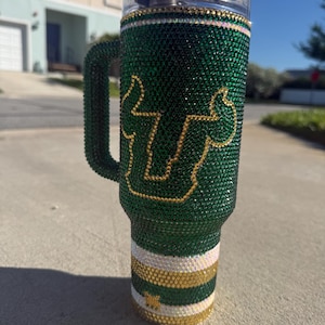 Puede incluir: Un vaso verde adornado con pedrer&iacute;a con asa, con un logotipo y detalles dorados. El vaso est&aacute; cubierto de brillantes pedrer&iacute;a verde, con rayas doradas y blancas cerca de la base. El asa tambi&eacute;n est&aacute; cubierta de pedrer&iacute;a verde.