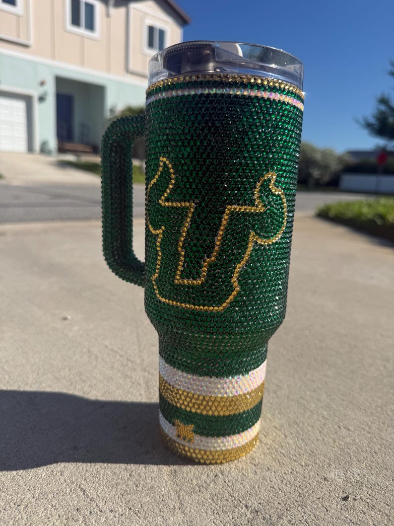 Puede incluir: Un vaso verde adornado con pedrer&iacute;a con asa, con un logotipo y detalles dorados. El vaso est&aacute; cubierto de brillantes pedrer&iacute;a verde, con rayas doradas y blancas cerca de la base. El asa tambi&eacute;n est&aacute; cubierta de pedrer&iacute;a verde.