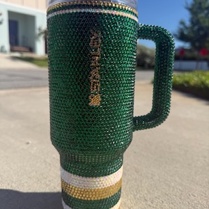 Puede incluir: Un vaso verde y dorado adornado con pedrer&iacute;a con asa. El vaso est&aacute; cubierto de pedrer&iacute;a verde, con rayas doradas y blancas en la base. El asa tambi&eacute;n est&aacute; cubierta de pedrer&iacute;a verde. El borde superior tiene pedrer&iacute;a dorada y transparente.