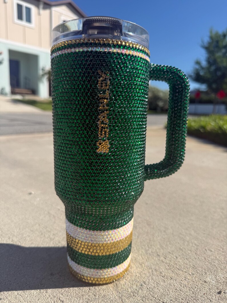 Puede incluir: Un vaso verde y dorado adornado con pedrer&iacute;a con asa. El vaso est&aacute; cubierto de pedrer&iacute;a verde, con rayas doradas y blancas en la base. El asa tambi&eacute;n est&aacute; cubierta de pedrer&iacute;a verde. El borde superior tiene pedrer&iacute;a dorada y transparente.