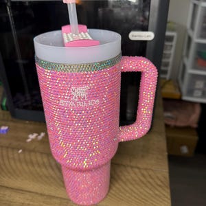 K&ouml;nnte beinhalten: Rosa, mit Strass besetzter Becher mit Henkel. Der Becher ist mit kleinen, schillernden Strasssteinen bedeckt. Ein wei&szlig;er Deckel mit rosa Schieber und ein dekorativer Strohhalm mit rosa Rose sind sichtbar. Auch der Griff ist mit Strasssteinen besetzt.