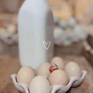 May include: A white ceramic milk bottle with a heart drawn on it stands behind a ceramic egg holder filled with six eggs. A small red flower is nestled among the eggs. The items are on a wooden surface.