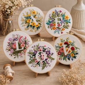 Könnte beinhalten: Fünf runde Stickrahmen aus Holz mit weißem Stoff, auf dem handgestickte Blumenkränze die Buchstaben A, E, L, O und V umrahmen. Jeder Rahmen zeigt farbenfrohe Stickereien mit verschiedenen Blumen, Blättern und Stängeln.