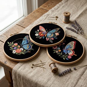 May include: Three embroidered butterfly hoops with wooden frames. Each features a black background with a colourful butterfly and floral design. The butterflies have intricate details in shades of blue, orange, and red. Sewing notions and lavender sprigs are also visible.