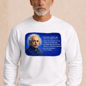 Puede incluir: Sudadera blanca de cuello redondo con un retrato de Albert Einstein y una cita sobre física en un rectángulo azul. La sudadera la lleva una persona con pelo gris y barba.