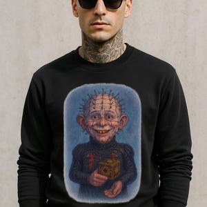 May include: Black sweatshirt featuring a portrait of a character with pins in their head, holding a puzzle box. The character has a sinister smile and is set against a blue background. The person in the image is wearing black sunglasses.