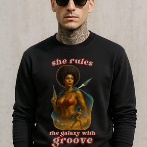 May include: Black sweatshirt featuring a retro illustration of a woman with a gun and a cape. The text "she rules the galaxy with groove" is printed in red. The person in the image is wearing sunglasses.