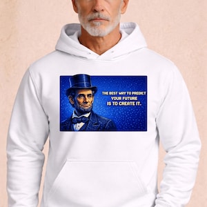 Puede incluir: Sudadera con capucha blanca con un gráfico de Abraham Lincoln y la cita: "LA MEJOR MANERA DE PREDECIR TU FUTURO ES CREARLO". La imagen de Lincoln está sobre un fondo azul. La sudadera tiene un bolsillo delantero y una capucha con cordón.