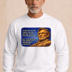 Puede incluir: Sudadera blanca con un retrato de Abraham Lincoln y una cita sobre la creencia. El texto de la sudadera dice: "Creer en las cosas que puedes ver y tocar no es creer en absoluto... Pero creer en lo invisible es un triunfo y una bendición."
