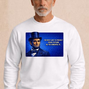 Puede incluir: Sudadera blanca con un gráfico rectangular de Abraham Lincoln con sombrero de copa, con la cita "LA MEJOR MANERA DE PREDECIR TU FUTURO ES CREARLO." La sudadera la lleva una persona con pelo gris y barba.