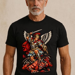 Könnte beinhalten: Schwarzes T-Shirt mit einem detaillierten Grafikdesign eines geflügelten Kriegers in Rüstung, der ein Schwert über einem roten Dämon hält. Der Krieger trägt einen roten Umhang und goldene Akzente.