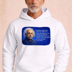 Puede incluir: Sudadera con capucha blanca con un retrato de Albert Einstein y una cita sobre la energía y la realidad en un rectángulo azul. La sudadera tiene un bolsillo de canguro y una capucha con cordón.