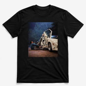 May include: A black short-sleeve t-shirt featuring a graphic print of a person lying on the ground with one leg propped up against the open door of a white sports car. The scene is set outdoors at night with trees visible in the background.