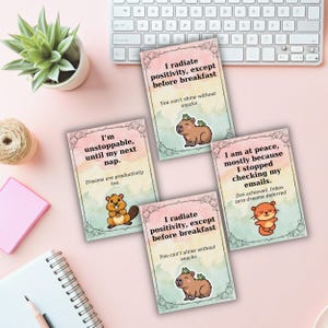 May include: Four cards with inspirational quotes and cute animal illustrations are arranged on a pink surface. The cards feature phrases like "I radiate positivity" and "I'm unstoppable." A keyboard and a potted plant are in the background.