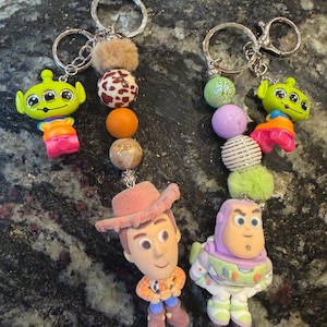 Pu&ograve; includere: Due portachiavi con personaggi del film Toy Story. Un portachiavi ha una figura di alieno verde, mentre l'altro ha una figura di Woody e Buzz Lightyear. Ogni portachiavi ha un anello argentato e un design di perline decorative con una palla soffice.