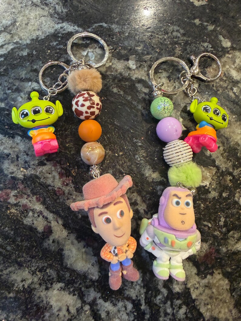 Pu&ograve; includere: Due portachiavi con personaggi del film Toy Story. Un portachiavi ha una figura di alieno verde, mentre l'altro ha una figura di Woody e Buzz Lightyear. Ogni portachiavi ha un anello argentato e un design di perline decorative con una palla soffice.