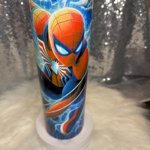 May include: A tall, cylindrical tumbler with a Spider-Man design. The artwork depicts the superhero in his red and blue costume, with blue lightning and a cloudy sky background. The tumbler is designed for drinks and is approximately 20 cm tall.