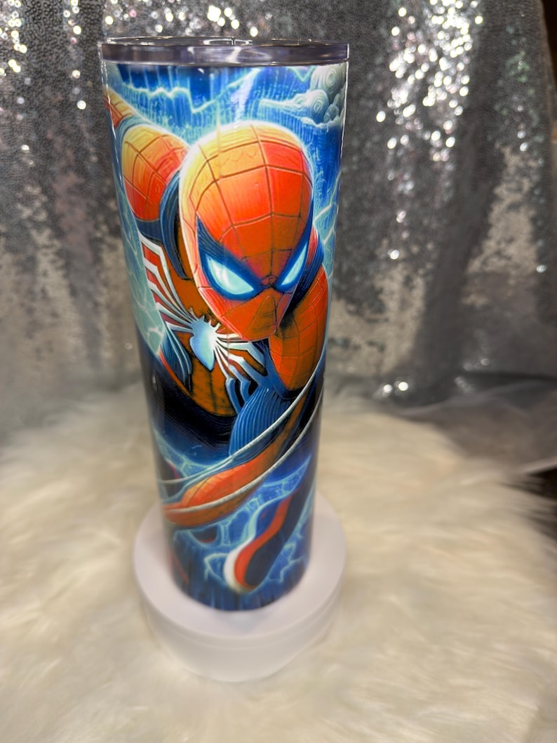 May include: A tall, cylindrical tumbler with a Spider-Man design. The artwork depicts the superhero in his red and blue costume, with blue lightning and a cloudy sky background. The tumbler is designed for drinks and is approximately 20 cm tall.