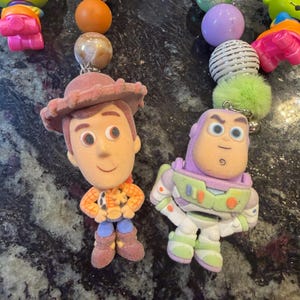 Pu&ograve; includere: Portachiavi con personaggi di Toy Story. I portachiavi includono Woody e Buzz Lightyear, insieme a perline e ciondoli colorati. Il portachiavi di Woody indossa un cappello marrone e il portachiavi di Buzz Lightyear indossa una tuta spaziale.