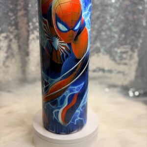 May include: A tall, cylindrical tumbler with a Spider-Man design. The tumbler features the superhero in his red and blue suit, set against a backdrop of blue lightning. The tumbler is likely made of stainless steel, and is approximately 20 cm tall.