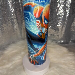 May include: A stainless steel tumbler with a colourful illustration of Spider-Man. The design shows the superhero in a dynamic pose with blue lightning effects and a cityscape backdrop. The tumbler is cylindrical and has a clear lid.