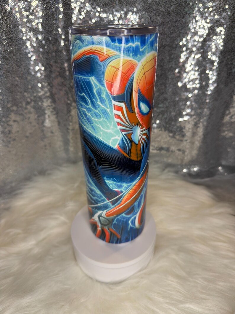 May include: A stainless steel tumbler with a colourful illustration of Spider-Man. The design shows the superhero in a dynamic pose with blue lightning effects and a cityscape backdrop. The tumbler is cylindrical and has a clear lid.