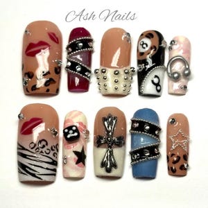 May include: A collection of ten artificial nails with diverse designs. The nails showcase lip, leopard print, and zebra stripe patterns, alongside studded, cross, and star decorations. Some nails have a nude base, while others are painted in shades of red, black, and blue.