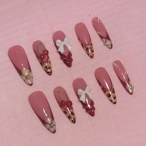 May include: A collection of ten long, almond-shaped artificial nails. The nails are painted with a pink base and decorated with white bows, cherry details, and leopard print tips. Some nails have a floral design.