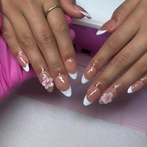 Peut inclure: Manucure française avec des ongles en amande. Les ongles ont une base transparente et des pointes blanches. Certains ongles sont décorés de petites fleurs roses. Une bague dorée est visible sur un doigt.