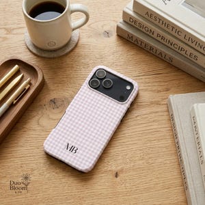 May include: A light pink and white gingham patterned smartphone case featuring the black monogram letters MB centered near the bottom. The case has a rectangular cutout for the camera lenses and is positioned on a light wood surface.