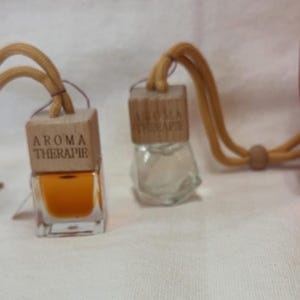 Pu&ograve; includere: Due piccole bottiglie di aromaterapia in vetro con tappi di legno e la scritta "AROMA THERAPIE". Una bottiglia contiene un liquido arancione. Le bottiglie sono collegate a un cordino marrone. Una zucca decorativa arancione &egrave; sullo sfondo.