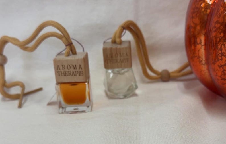 Pu&ograve; includere: Due piccole bottiglie di aromaterapia in vetro con tappi di legno e la scritta "AROMA THERAPIE". Una bottiglia contiene un liquido arancione. Le bottiglie sono collegate a un cordino marrone. Una zucca decorativa arancione &egrave; sullo sfondo.