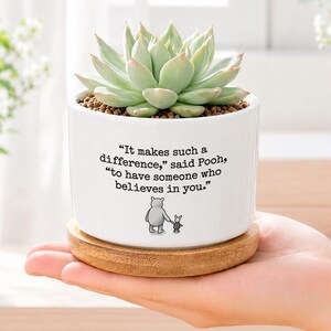 Può includere: Un vaso cilindrico in ceramica bianca contenente una pianta succulenta verde. Il vaso presenta una citazione stampata in nero che recita: "It makes such a difference," said Pooh, "to have someone who believes in you." e una piccola illustrazione di Pooh e Pimpi.