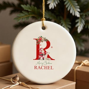May include: A round, off-white Christmas ornament with a gold string. The ornament features a red letter "R" with a Santa Claus illustration and the text "Merry Christmas RACHEL".