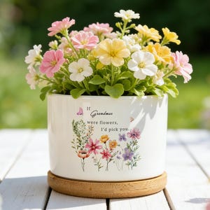 Puede incluir: Una maceta de cerámica blanca llena de flores coloridas en tonos rosa, amarillo y blanco. La maceta tiene un diseño floral y el texto "If grandmas were flowers, I'd pick you." Se encuentra sobre un posavasos redondo de madera.