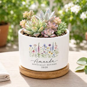 Peut inclure: Un pot de fleurs en céramique blanche contenant diverses plantes succulentes, posé sur une base en bois circulaire. Le pot présente une illustration de fleurs sauvages et le texte imprimé : Amanda, OFFICIALLY RETIRED, 2026.